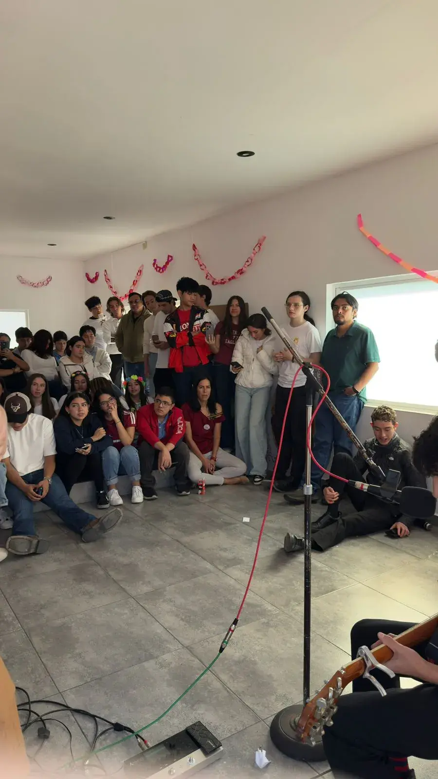 Grupo numeroso de jóvenes en un salón decorado, algunos sentados y otros de pie, con una persona tocando guitarra y micrófono con cables visibles.