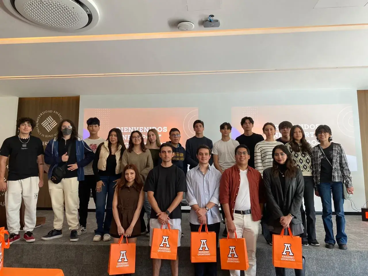 Grupo de jóvenes posando para una foto en un evento educativo interior con pantallas que muestran un mensaje de bienvenida y bolsas de regalo naranjas visibles.
