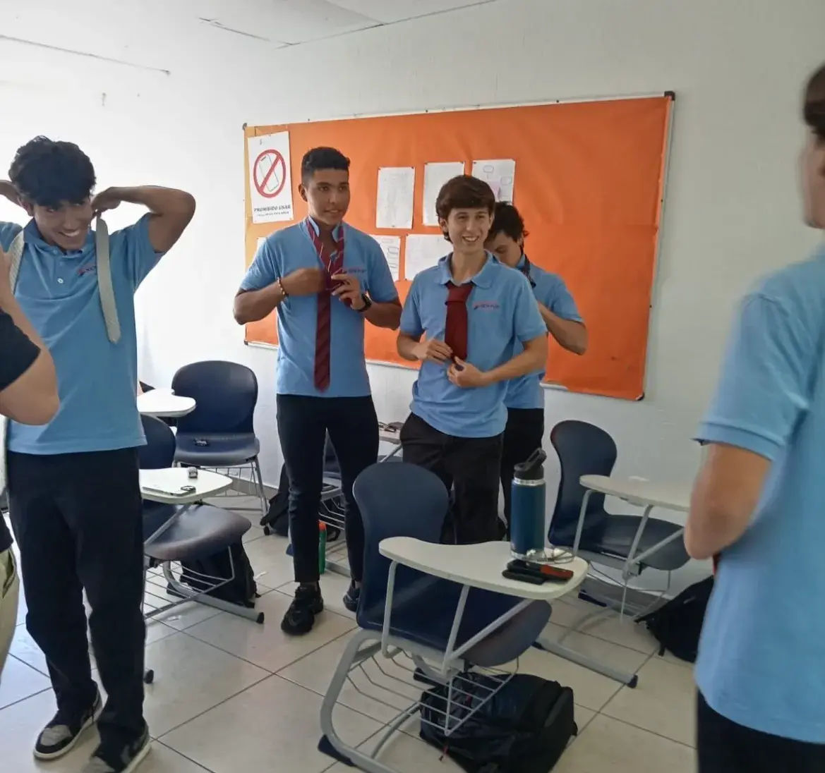 Grupo de estudiantes en aula de New Port High School con camisas azules y pantalones oscuros, ajustando corbatas, frente a sillas, escritorios y un tablón naranja.