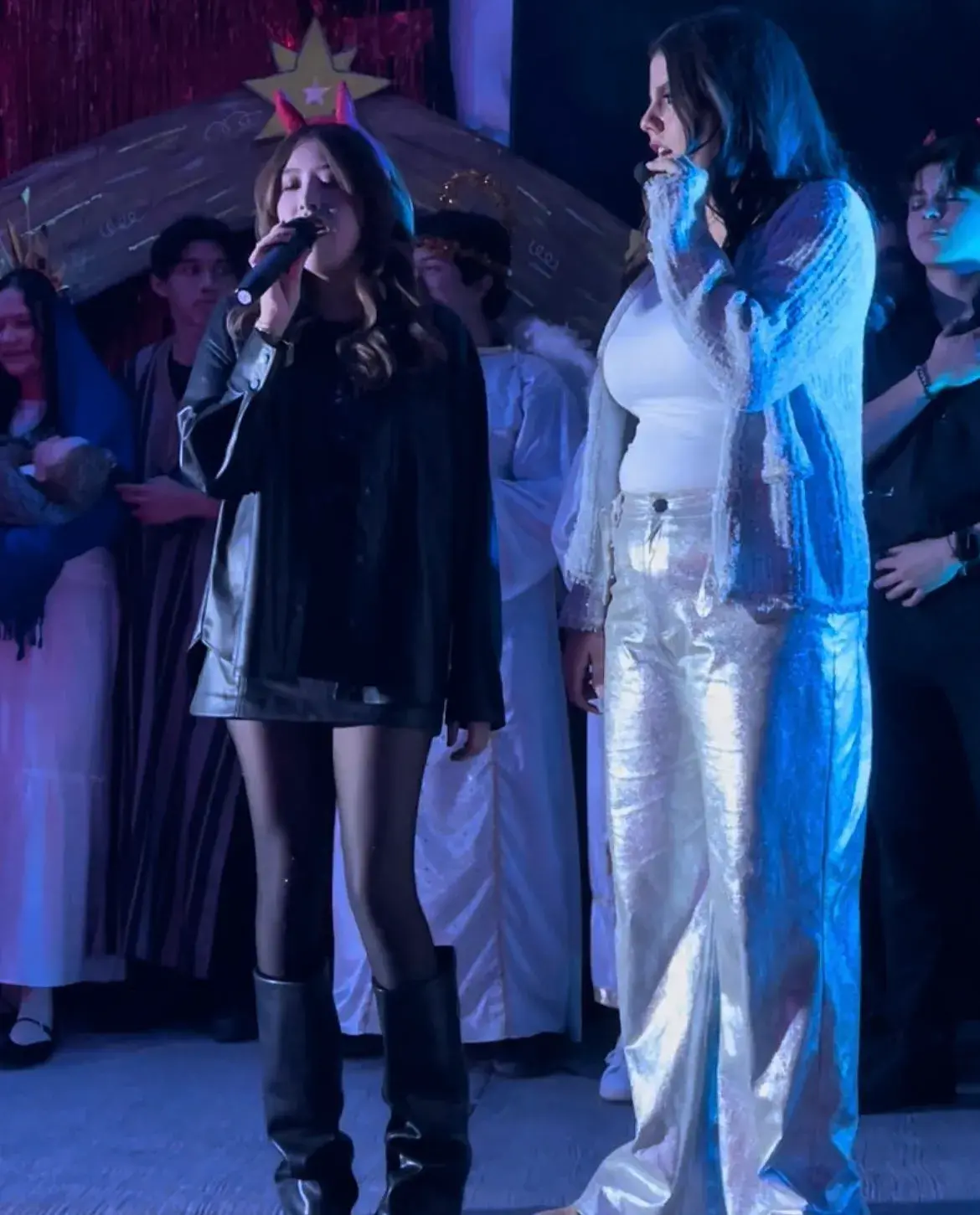 Personas en escena realizando una presentación artística, una mujer canta con micrófono y otra está al frente, ambas acompañadas por personas en disfraces de ángeles y otros roles.