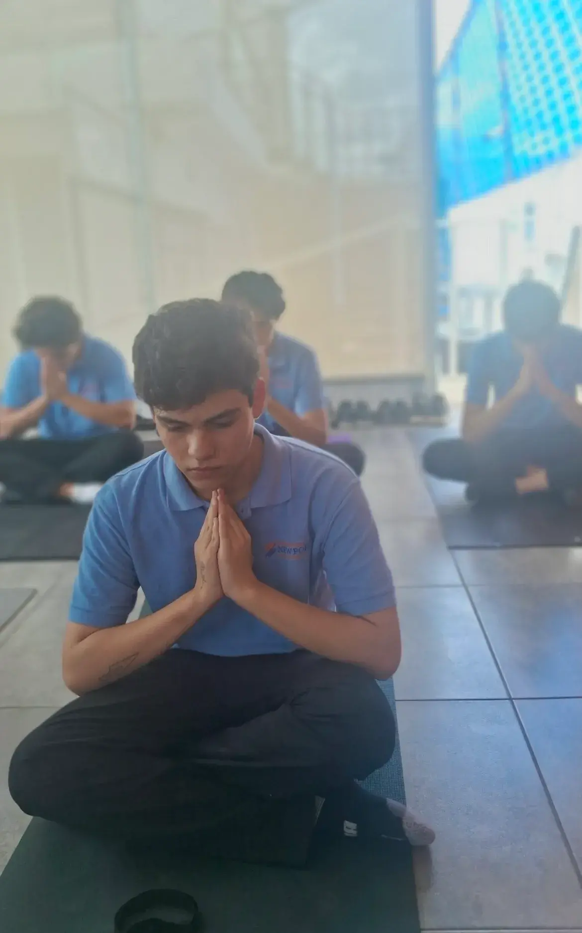 Group of young men sitting cross-legged on yoga mats indoors with hands clasped in meditation.