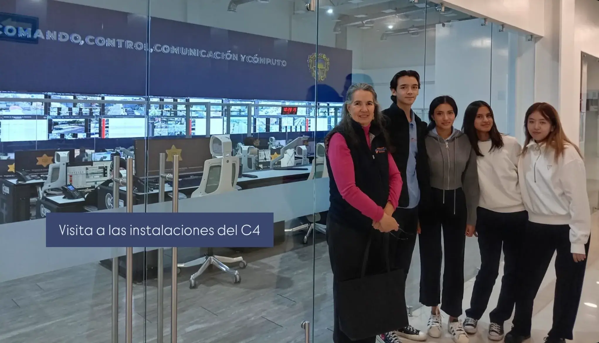 Five people standing in front of a glass wall showing a control room with computer monitors and control panels inside