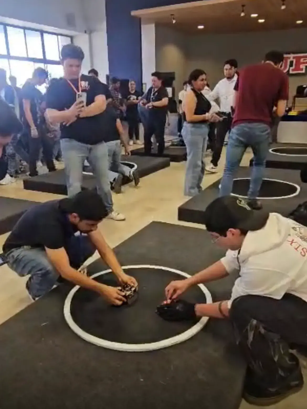 Indoor scene of a robotics competition with participants operating small robots inside circular arenas on raised platforms, some using smartphones to photograph.