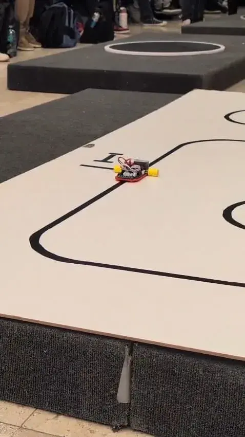 Robot pequeño avanzando por una pista con líneas negras mientras personas observan de pie al fondo